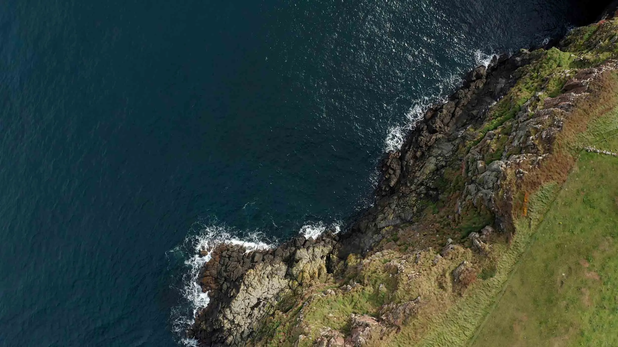An aerial view of dark blue waves crashing against the grassy cliff at Rathlin.